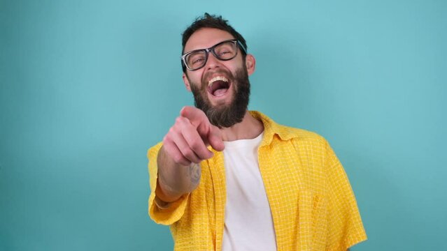 A bearded hipster laughs out loud pointing his finger at the camera, making fun of other people's failures. The guy laughs at his friend's good joke.