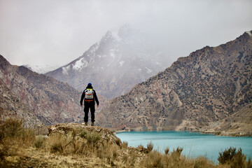 A man with a backpack stands on a mountainside and looks into the distance at a mountain lake and peaks. A lone hiker travels high in the mountains on a cloudy day. Independent hike.
