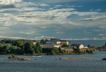 Helsinki, Finland - July 20, 2022: Suomenlinna Fortress from the sea. Ramparts and yellow buildings and terrain of the Naval Academy o Pikku Musta island under blue cloudscape. Islets in sea