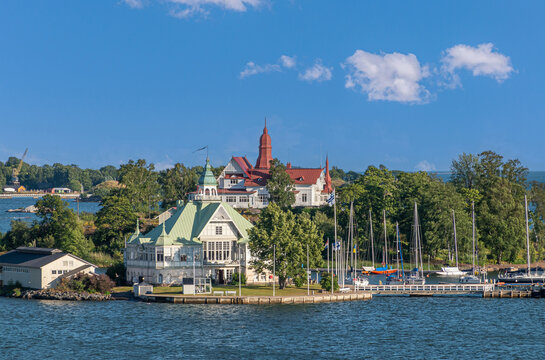 Helsinki, Finland - July 19, 2022: Closeup, Ravintola NLK on its Valkosaari island had green roof. Restaurant Saaristo onn its island has red roof under blue cloudscape. Yachts in port 