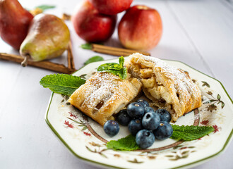 A traditional piece of apple strudel with powdered sugar, mint, blueberry and cinnamon in the foreground on a table.