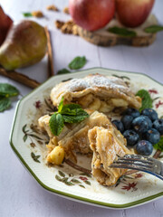 A traditional piece of apple strudel with powdered sugar, mint, blueberry and cinnamon in the foreground on a table.