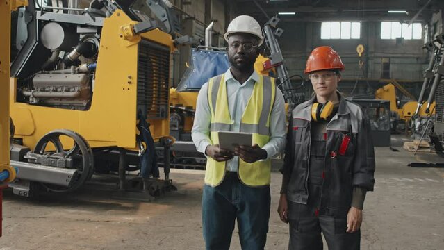 Medium portrait of African American male engineer with tablet computer and Caucasian female plant worker in coverall posing for camera standing at tractor plant