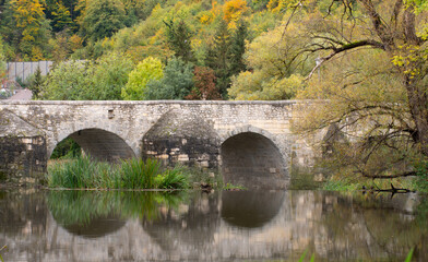 Fototapeta na wymiar A historic stone bridge, with several round arches, is reflected in the water. In the background deciduous trees in autumn with golden leaves.