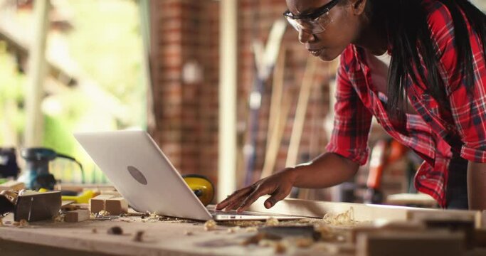 African American female carpenter working with computer laptop in workshop . craftsman wearing safety glasses checking order of clients or learning online