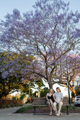 Obraz premium lesbian couple sitting on park bench under beautiful purple tree