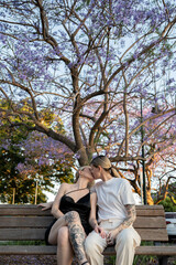 lesbian couple sitting on park bench intimate kissing under beautiful purple tree 