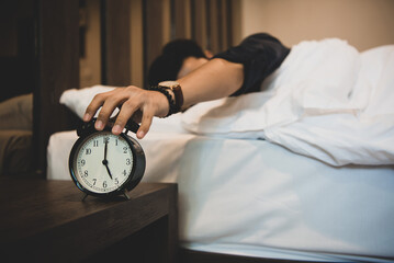 A young man is lying on the bad at home, he is sliding the alarm clock with his hand placed at the bedside table. He pressed the alarm clock out in the morning so he could continue sleeping.