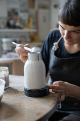 Focused woman in apron restores ceramic vessel found during archaeological excavations by treating it protective liquid. Professional lady in pottery workshop makes dishes for sale at online auctions