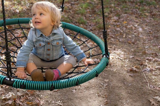 Blond two year old sitting on a rope swing outdoors; hipster baby in jean jacket