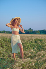 stylish beautiful smiling young girl in a straw hat and a fashionable dress walks around the field and poses