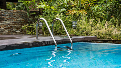 A staircase with a metal railing descends into the swimming pool. There are flip-flops nearby. Highlights on turquoise water. There is lush tropical vegetation nearby.