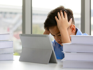 Young Asian elementary school boy sitting next to stack of books and using tablet computer with both hands holding his head with gestures and face showing stress. - 545336183