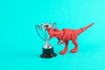 Toy red dinosaur tyrannosaurus rex with winner cup on turquoise background. Minimalism creative...