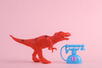 Toy red dinosaur tyrannosaurus rex with retro rotary phone on pink background. Minimalism creative...