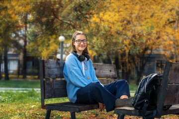 A young schoolgirl with a backpack and headphones is sitting on a bench on a sunny day.