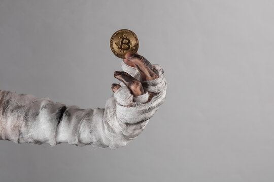 Mummy's hand wrapped in bandage holds bitcoin isolated on gray background. Halloween, business concept