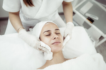 Obraz na płótnie Canvas Facial massage and skincare treatment. Dermatologist hands cleaning relaxed serene young woman face with napkin in beauty salon during skincare treatment.