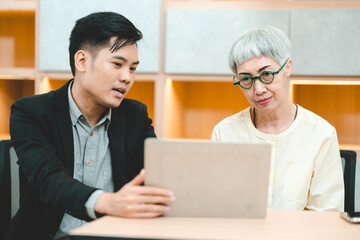 Young Asian business man in formal clothing showing something on digital tablet to senior female boss and manager wearing eyeglasses while in a meeting and training on technology