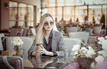 Young blonde woman in sunglasses holds a cup of coffee while sitting at a table in the summer terrace of cafe