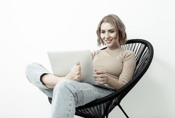 Young smiling woman looks into the laptop screen while sitting on a comfortable armchair barefoot. Lifestyle