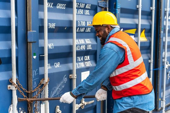 Photo of a black african engineer using a crow bar to force open the chain on a container in a shipping containers yard