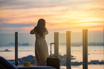 Beautiful woman in dress with suitcase on vacation by tropical beach, standing on the glass balcony enjoy fresha air with ocean view. Golden sunset sky on the beach, Beautiful cloudscape over the sea.