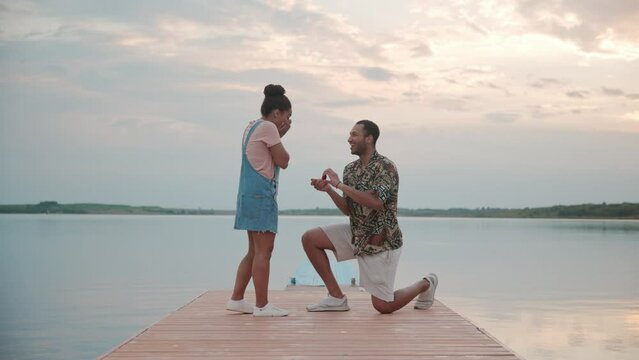 Young handsome man standing on one knee making proposal to his beloved woman giving her engagement ring on lake. Happy girlfriend take a ring accepting proposal of marriage. Just engaged, love concept