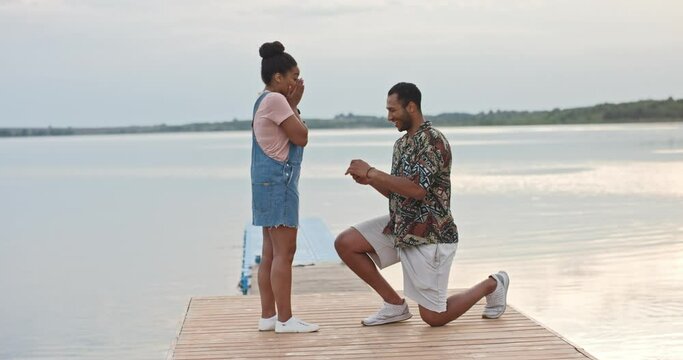 Young cheerful handsome man standing on one knee making proposal of marriage to his beloved woman on dock on lake giving engagement ring. Happy surprised woman got engaged with boyfriend. Love concept
