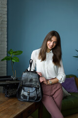 beautiful slender smiling girl in burgundy pants and a white blouse posing with a black leather backpack