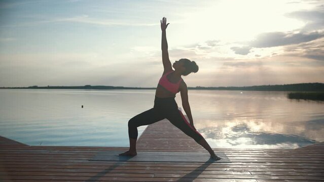 One African American young sporty sportswoman in sportswear practicing yoga on lake on pier on sunrise or sunset. Pretty woman athlete having workout on nature standing in yoga warrior posture