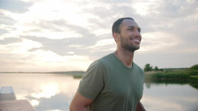 Close up portrait of joyful handsome young sportsman running on nature outdoors in good mood and smiling. Morning workout. African American attractive male athlete runs on dock on lake on sunset