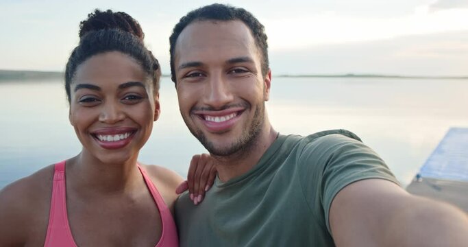 Close up joyful young couple in love man and woman standing on lake posing to smartphone camera smiling and hugging after workout. POV African American family couple taking selfie photo on cellphone