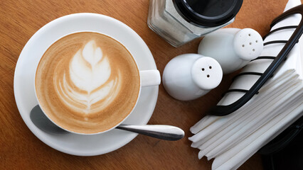 Delicious golden flat white coffee with flower art in white cup and saucer on light wooden table, birds eye topdown in local coffee shop