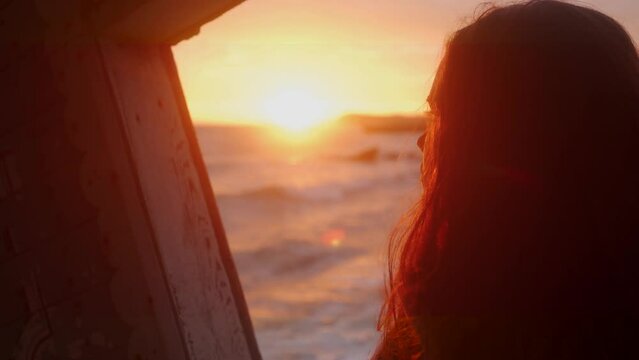 Woman stands in old wooden ruined leaning house interior on sea beach, looks at window on sunset. Mixed ethnic female enjoys view from vintage abandoned rickety home on coastline. Dreamy, enigmatic.
