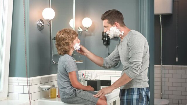Close up of little teen boy sitting on sink indoor in bathroom listening to father how to shave face with razor. Caucasian dad teaching his kid boy to shave using shaving cream and razor. Self care
