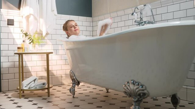 Beautiful young woman taking a bath indoors at home. Happy pretty lady bathing with foam. Cheerful female in tub washing body with soap relaxing in modern bathroom. Personal hygiene.