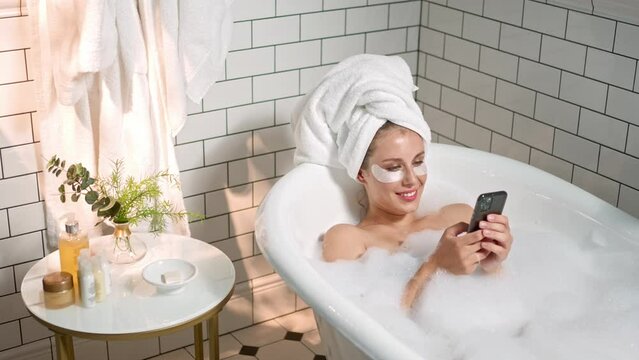 Top view of Caucasian attractive woman with towel on head lying in relaxing bath with soap bubbles smiling and typing on smartphone browsing on social media. Leisure. Young lady using cellphone in tub