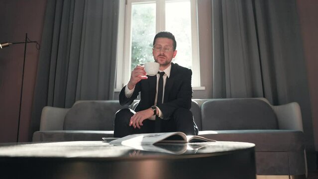 Handsome stylish elegant businessman in suit sitting on sofa in hotel room texting on smartphone and drinking coffee while waiting. Caucasian young adult confident male indoor in luxury lobby