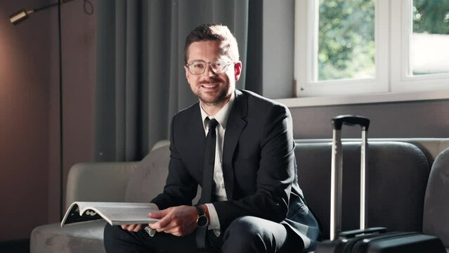 Portrait of happy successful businessman sitting on sofa in luxury hotel reading journal while waiting for business meeting and smiling to camera. Elegant male reading magazine indoors. Business trip