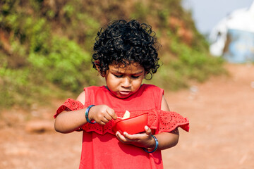 Adorable child with curly hairs eating fruits with chopstick ,holding bowl in hand.