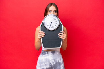 Young beautiful caucasian woman isolated on red background with weighing machine and hiding behind it