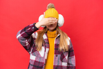 Young beautiful woman wearing winter muffs isolated on red background covering eyes by hands. Do...