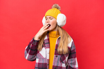 Young beautiful woman wearing winter muffs isolated on red background yawning and covering wide open mouth with hand