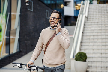 A smiling casual businessman is pushing his bicycle on the street and talking on the phone.