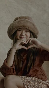 Young asian girl standing in front of a backdrop in photography studio looking confident, showing heart shape with hands