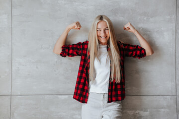 Excited caucasian blonde woman in plaid shirt shows double biceps against marble wall, satisfied by her health. Fitness concept. Cheerful model at studio. Pretty housewife at home. Strength concept.