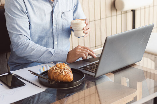Man typing on laptop and holding coffee cup with snacks next to it - Powered by Adobe