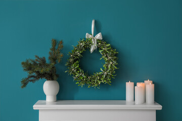 Hanging Christmas mistletoe wreath, vase with fir branches and candles on mantelpiece near blue wall