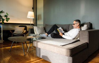 Middle age man with eye glasses reading book at home on couch.